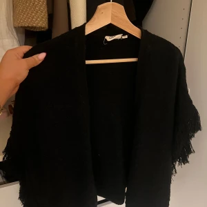 Svart poncho med fransar - Säljer en snygg svart poncho från Kappahl. Den är perfekt för höst och vår, med fransar längs kanterna som ger en bohemisk touch. 