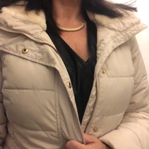 Beige dunjacka med pälskantad huva - Säljer en supermysig beige dunjacka med pälskantad huva. Jackan har dragkedja och knappar framtill, samt praktiska fickor med dragkedja. Perfekt för kalla vinterdagar! 🧥❄️
