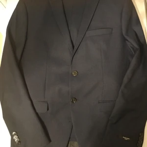 Mörkblå kostym från Jack & Jones - Säljer en stilren mörkblå kostym storlek 50 från Jack & Jones i super slim passform. Kavajen är i polyester och har snygga knappar framtill. Perfekt för både fest och formella tillfällen. Tillverkad i Turkiet och i mycket bra skick.