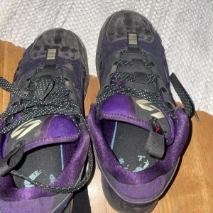 Svarta och lila sneakers - Säljer ett par coola svarta sneakers med lila detaljer och snörning. De har en unik design med turkosa stänk på sidorna och en slitstark sula. Perfekta för vardagsbruk eller träning. Skorna är i bra skick men har lite slitage på sulan.