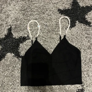 Svart ribbad topp från FB Sister - Säljer en svart ribbad topp från FB Sister i storlek XXS. Toppen har justerbara axelband dekorerade med pärlor och en söt fjärilsdetalj. Perfekt för en somrig look eller en utekväll. Toppen är i mycket bra skick och redo för nya äventyr!