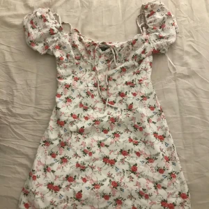 Blommig sommarklänning med puffärmar - Söt blommig klänning i vitt med röda och gröna detaljer. Den har puffiga kortärmar och en knytning framtill. Fick i present men passar tyvärr inte
