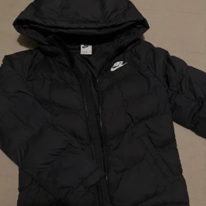 Svart dunjacka från Nike - Säljer en svart dunjacka från Nike i storlek S för barn (128-137 cm). Jackan har en huva och dragkedja framtill, perfekt för kyliga dagar. Den är i mycket bra skick och passar både höst och vinter. En stilren och bekväm jacka för alla tillfällen!