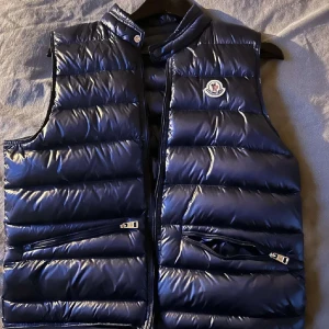 Mörkblå dunväst från Moncler - Säljer en snygg och stilren mörkblå dunväst från Moncler. Västen har en glansig finish och är perfekt för kyliga dagar. Den har dragkedja framtill och två praktiska fickor med dragkedjor. Moncler-loggan sitter på bröstet. Perfekt för höst och vinter! Size xs Recomenderar att vara 160-170. Pris kan diskuteras 