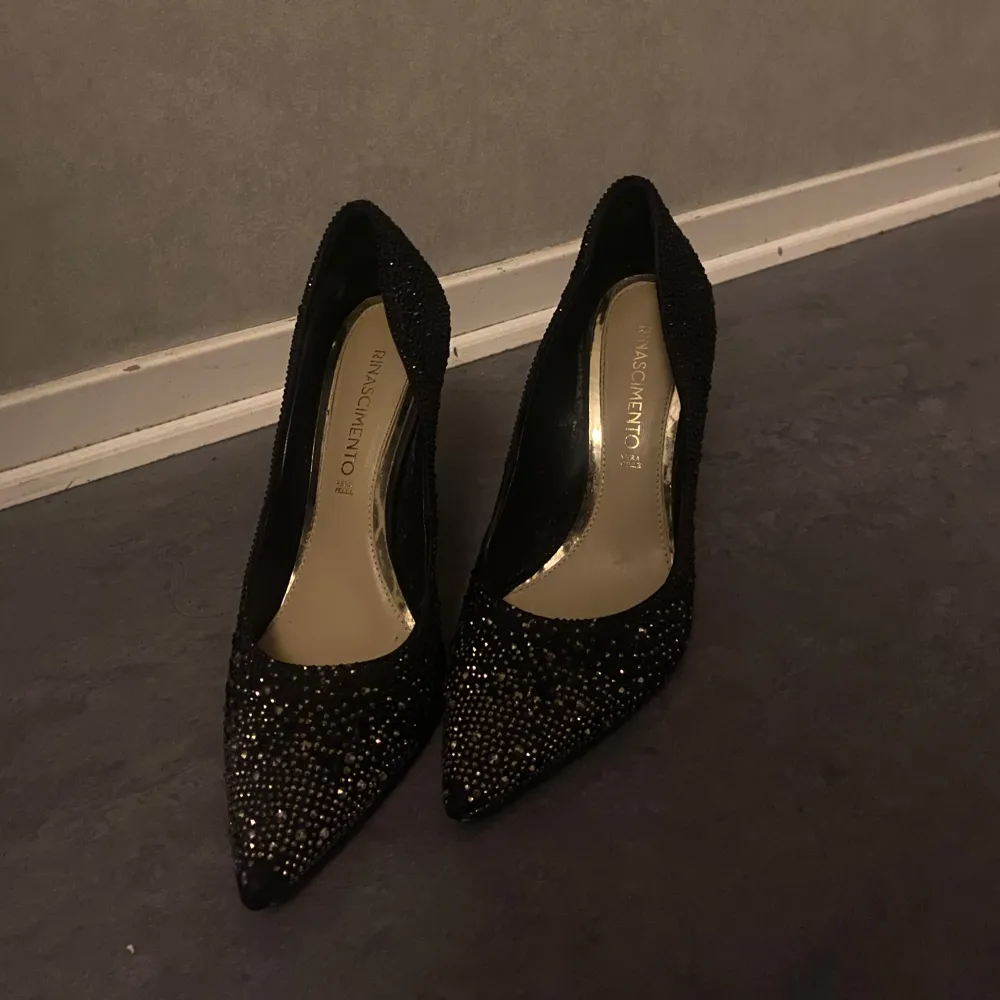 Snygga svarta klackar från rinascimento i glittrigt material. De har en elegant spetsig tå och en hög klack som ger en stilren look. Perfekta för festliga tillfällen när du vill glänsa lite extra. Använt dem 1-7 gånger köpta för 1900 på en italiensk butik . Kengät.