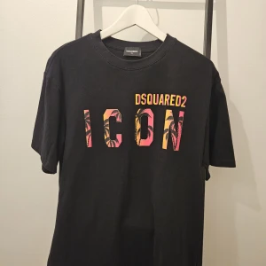 Svart t-shirt från Dsquared2 - Säljer en svart t-shirt från Dsquared2 i storlek XL. Den har en cool text 'ICON' i rosa och orange med palmträdsmönster. Perfekt för en avslappnad stil eller fest. T-shirten är i mycket bra skick och gjord av mjukt material. OBS LITEN I STORLEK SIZE SITTER SOM STORLEK L