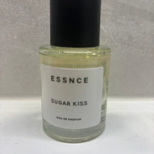 Essnce Sugar Kiss Eau de Parfum - Säljer en flaska Essnce Sugar Kiss Eau de Parfum. Den har en söt och fräsch doft som passar perfekt för vardagsbruk. Flaskan är stilren och enkel med en svart kork och en vit etikett. Perfekt för dig som älskar en söt och lätt doft!