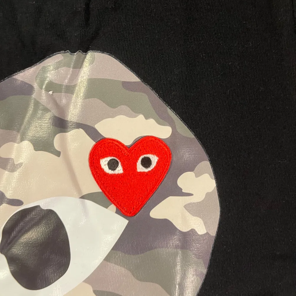 Säljer en svart t-shirt från Comme des Garçons med ett stort hjärta i camouflage på framsidan. T-shirten är kortärmad och i bomull, perfekt för en avslappnad stil. Den har en cool och unik design med ett rött hjärta inuti det stora hjärtat. Passar perfekt till både vardag och fest! Säljer för 699 . T-shirts.