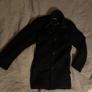 Marinblå kappa från Indigo - Säljer en stilren marinblå kappa från Indigo i storlek L. Jackan har både dragkedja och knappar framtill, vilket ger extra skydd mot kyla. Perfekt för höst och vinter med sin långa ärm och klassiska design. Nypris 1850kr, Vårat pris 299kr