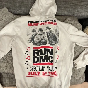 Vit RUN DMC hoodie från H&M - Säljer en vit hoodie från H&M med coolt RUN DMC-tryck. Perfekt för dig som älskar retro och hiphop! Den har en stor bild på baksidan och en mindre logga fram. Grym för både vardag och chill. 🕶️