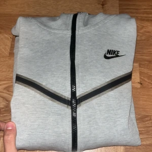 Grå hoodie från Nike - Säljer en grå hoodie från Nike i storlek S. Den har en dragkedja framtill och en klassisk Nike-logga på bröstet. Perfekt för höst och vår, med en bekväm passform och huva. Gjord i mjukt material för extra komfort.