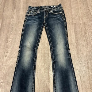 Jeans med broderade detaljer från Miss Me - Snygga jeans från Miss Me i mörkblå denim med coola broderade detaljer längs sidorna och på bakfickorna. De har en bootcut-stil och är perfekt för en avslappnad men ändå stilren look. Jeansen har också en unik sliten effekt som ger dem en extra edge.