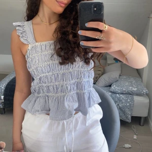 Blå- och vitrandig topp med volang - Söt blå- och vitrandig topp med volangdetaljer och smockad design. Den är ärmlös med breda axelband och har en luftig passform, perfekt för sommardagar. Toppen är både stilren och bekväm, idealisk för en avslappnad look.