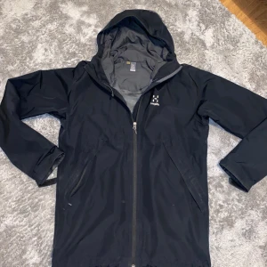 Svart regnjacka från Haglöfs - Säljer en svart regnjacka från Haglöfs i bra skick. Den har en praktisk huva och dragkedja framtill. Perfekt för regniga dagar och håller dig torr och bekväm. Passar både till vardags och utomhusaktiviteter. Hör av dig vid intresse! Den är dock riven vid fickan 