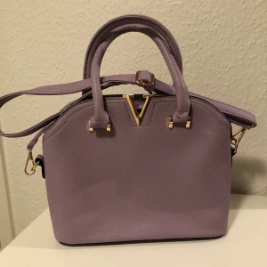 Ljuslila handväska med gulddetaljer - Säljer en superfin ljuslila handväska i syntetmaterial med snygga gulddetaljer. Väskan har både handtag och en avtagbar axelrem, vilket gör den perfekt för både vardag och fest. Den har en stilren design med en V-formad gulddetalj på framsidan. Väskan är i mycket bra skick och redo för nya äventyr!