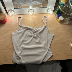 Vit ribbad linne med spetsdetaljer - Säljer ett supergulligt vitt ribbat linne med spetsdetaljer vid axelbanden och längs halsringningen. Perfekt för sommardagar eller som ett lager under en tröja. Linnet har en söt rynkning framtill som ger det lite extra stil. Passar perfekt för en avslappnad look eller en dag på stan.