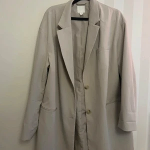 Beige kappa från H&M - Säljer en stilren beige kappa från H&M i storlek M. Den har en klassisk design med knappar framtill och långa ärmar. Perfekt för höst och vår, och ger en elegant touch till vilken outfit som helst.