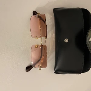 Stilrena solglasögon med rosa tonade glas - Säljer ett par snygga solglasögon med rosa tonade glas och guldfärgade detaljer. De har en rektangulär form och kommer med ett svart fodral. Perfekta för en solig dag och ger en touch av glamour till din outfit. Passar både till vardags och fest!