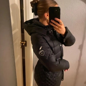 Svart dunjacka från Rockandblue - Säljer en snygg svart dunjacka från Rockandblue i storlek 38. Jackan har en quiltad design med dragkedja och knappar framtill. Perfekt för kalla vinterdagar med sin varma och stilrena look. Den har en huva och flera praktiska fickor med dragkedjor. Man kan få till en fakepäls luva till.