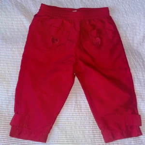 Vinröda byxor med hjärtdetaljer - Vinröda barn fodrade sommarbyxor med raka ben, i storlek 74 från Lindex. Söta röda byxor för barn med hjärtdetaljer på fickorna. Byxorna har en elastisk midja för bekväm passform och justerbara benslut med kardborreband. Perfekta för lek och vardag!