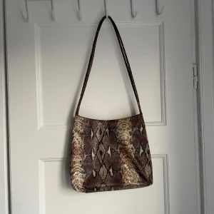 Ormmönstrad axelväska - Säljer en supercool axelväska med ormmönster i bruna och beige toner. Väskan har en dragkedja upptill och är perfekt för att ge din outfit en edgy touch. Den är i bra skick och redo för nya äventyr!