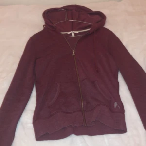 Vinröd hoodie från Victoria's Secret - Säljer en superfin vinröd hoodie från Victoria's Secret i storlek XS. Den har en dragkedja med en söt ängelvinge-detalj och matchande tryck på fickan, hoodien har också ängel vingar på ryggen av silvriga paljetter. Perfekt för mysiga höstkvällar eller som ett lager under jackan eller på en dag när det känns lite kyligare, de dagarna är de skönt att ta på sig något som håller en varm men endå som är super snyggt. Skriv till mig om ni har mer frågor