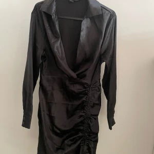 Svart långärmad klänning - Elegant svart klänning med långa ärmar och djup v-ringning. Klänningen har en stilren design med rynkade detaljer på sidan som ger en smickrande passform. Perfekt för en kväll ute eller en speciell tillställning.