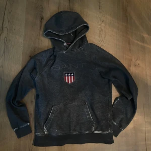 Grå hoodie från GANT - Säljer en snygg grå hoodie från GANT med ett broderat emblem på bröstet. Tröjan har en klassisk känguruficka och en bekväm huva. Perfekt för en avslappnad stil.