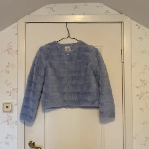Ljusblå teddyjacka från Lindex Kids - Mysig ljusblå jacka från Lindex Kids i storlek 146/152. Jackan har en fluffig textur och långa ärmar. Den är både stilren och bekväm, vilket gör den till ett måste i garderoben.