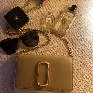 Beige axelväska med guldkedja - Snygg beige axelväska från Marc Jacobs med guldkedja och stort guldfärgat spänne på framsidan. Väskan har flera fack inuti, inklusive kortfack med rosa detaljer. Perfekt för att bära det viktigaste med stil.