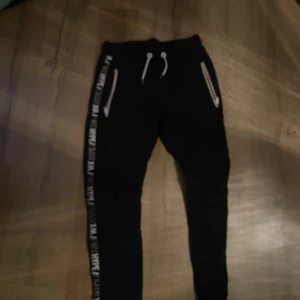 Svarta mjukisbyxor med textdetaljer - Svarta mjukisbyxor från Woxo med vita textdetaljer längs sidorna. Byxorna har snörning i midjan och dragkedjor vid fickorna för en sportig look. Perfekta för en avslappnad stil.