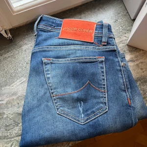 Blå jeans från Jacob Cohen BARD - Snygga blå jeans från Jacob Cohen med orangea sömmar och en unik design. De har en klassisk femficksmodell och en knappgylf. Perfekta för en stilren look. Slitningarna är design. Storlek 30, ställ frågor i DM, nypris runt 5000kr. Modellen BARD vilket är en nyare modell version som dem flesta inte har. 