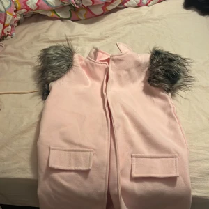 Rosa väst med pälsdetaljer - Säljer en söt rosa väst med grå pälsdetaljer vid axlarna. Västen har två fickor framtill och en stilren design utan knappar eller dragkedja. Perfekt för att lägga till en touch av stil till din outfit. Denna kappan är till barn som är 9-10 år verkligen jätte söt 