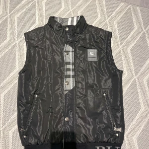 Svart quiltad väst från Burberry - Snygg svart quiltad väst från Burberry med klassiskt rutigt mönster på baksidan. Västen har dragkedja och knappar framtill samt ribbad kant med Burberry-logga. Perfekt för att hålla stilen i kyligare väder.