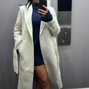 Vit kappa - Säljer en stilren vit kappa med klassisk design. Den har långa ärmar och en elegant krage. Perfekt för att hålla sig varm och snygg under kyligare dagar. Passar bra till både vardag och mer uppklädda tillfällen.