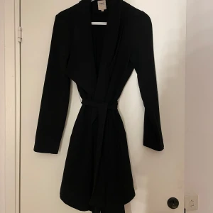 Svart kappa från ONLY - Snygg svart kappa från ONLY i storlek S. Den har en elegant design med ett bälte i midjan för en smickrande passform. Perfekt för höst och vår med sina långa ärmar och stilrena look.