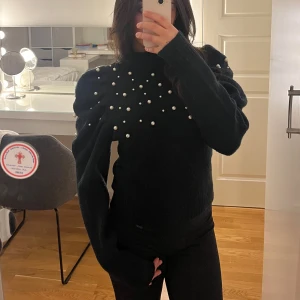 Svart stickad tröja med pärlor - Säljer en elegant svart stickad tröja med pärldetaljer över bröstet och ärmarna. Tröjan har en hög krage och långa ärmar med puffiga axlar som ger en trendig look. Perfekt för att ge din outfit det lilla extra. 🖤