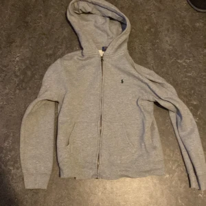 Grå hoodie från Ralph Lauren (152 cm) - lSäljer en stilren grå hoodie från Ralph Lauren med dragkedja och klassisk logga på bröstet. liten i storleken och ganska tvättads ungefär för en 12 åring. Perfekt för en avslappnad look. Den har en bekväm passform och är tillverkad i mjukt material.