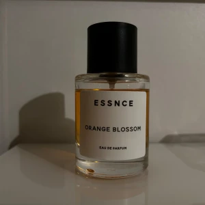 Essnce Orange Blossom Eau de Parfum - Upptäck Essnce Orange Blossom Eau de Parfum med en fräsch och blommig doft. Denna parfym har hjärtnoter av kaprifol, ros, apelsinblomma, iris och jasmin. Flaskan är elegant med en enkel design och svart kork.