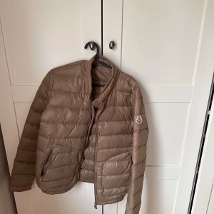 Beige dunjacka från Moncler - Säljer en snygg beige dunjacka från Moncler. Jackan har en stilren design med dragkedja och Moncler-logga på ärmen. Perfekt för kyligare dagar med sin lätta och värmande dunstoppning.
