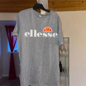 Grå t-shirt från Ellesse - Säljer en grå t-shirt från Ellesse med deras ikoniska logga i vitt, rött och orange på bröstet. T-shirten har en klassisk rund hals och korta ärmar. Perfekt för en avslappnad stil. Har en fläck på det vita texten som tyvärr inte går bort i tvätten
