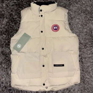 Canada goose väst - Säljer en stilren vit dunväst från Canada Goose med klassisk logga på bröstet. Västen har knappar och dragkedja framtill samt två praktiska fickor. Perfekt för kyliga dagar.