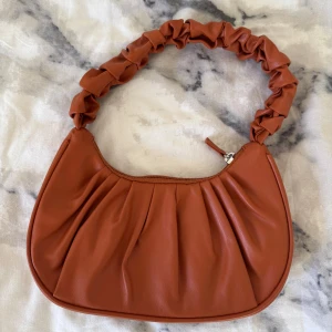 Brun axelväska med rynkad rem - Snygg brun axelväska i syntetmaterial med en unik rynkad rem. Väskan har en dragkedja upptill och en stilren design som passar perfekt för en trendig look. Perfekt för att bära dina nödvändigheter med stil. 