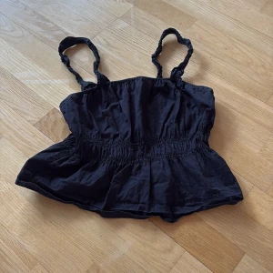 Svart topp - Säljer en svart topp med smockdetaljer och volang nedtill. Perfekt för varma sommardagar eller som ett lager under en jacka. Toppen har en luftig och bekväm passform.