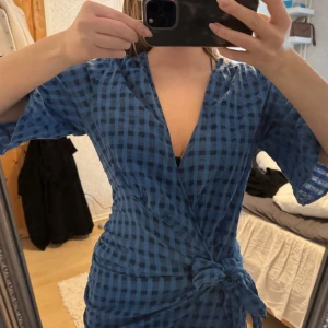 Blå rutig omlottklänning - Säljer en snygg blå rutig omlottklänning med trekvartsärmar och knytning i midjan. Klänningen har en v-ringad design och är perfekt för en stilren look.