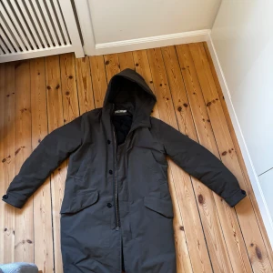 Skogsgrön parkajacka från Hope - Säljer en stilren brun parkajacka från Hope. Jackan har en praktisk huva och stängs med både dragkedja och knappar. Perfekt för kyligare dagar med sin långa design och quiltade insida.