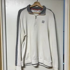 Vit och grå tröja från Calvin Klein - Snygg vit tröja med grå detaljer från Calvin Klein. Tröjan har en dragkedja vid halsen och ett CK-märke på bröstet. Perfekt för en stilren och avslappnad look.