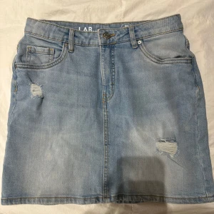 Ljusblå jeanskjol från LAB Industries - Trendig ljusblå jeanskjol från LAB Industries med slitna detaljer och klassisk femficksdesign. Perfekt för en avslappnad stil. Tillverkad av hållbart denim.