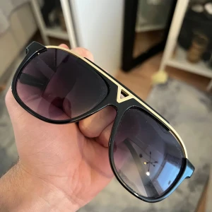 Lv glasögon  - Snygga svarta solglasögon med gulddetaljer och mörka linser. De har en klassisk design med en modern twist, perfekt för att ge en stilren look. Ramen är robust och har en elegant guldfärgad kant som ger en lyxig känsla.