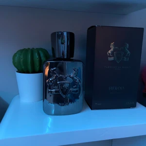 Parfym Herod från Parfums de Marly - Elegant parfymflaska i silver med en stilren design och ett emblem av två hästar. Flaskan rymmer 75 ml av doften Herod, känd för sina rika och kryddiga noter. Perfekt för den som söker en sofistikerad och tidlös doft.